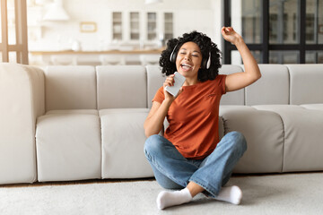 Happy black woman listening music and singing, sitting on floor at home