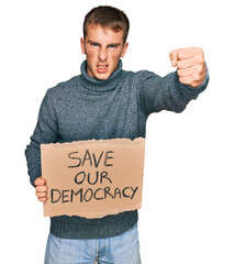 Young blond man holding save our democracy protest banner annoyed and frustrated shouting with...