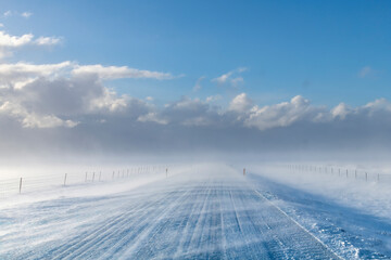 View over snow covered road in agricultural landscape in Iceland with high winds blowing snow over road creating a blur with snow clouds in distance giving way to clear blue sunny sky - obrazy, fototapety, plakaty