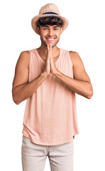 Young hispanic man wearing summer hat praying with hands together asking for forgiveness smiling confident.