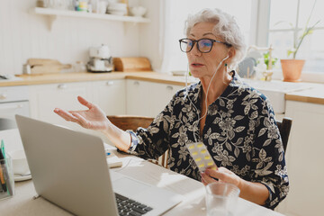 Senior female consulting with doctor sitting at kitchen table in front of laptop in earphones,...