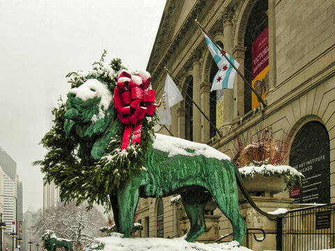 A bronze lion statue wearing a festive wreath with a red bow stands in front of the snow-covered Art Institute of Chicago building.