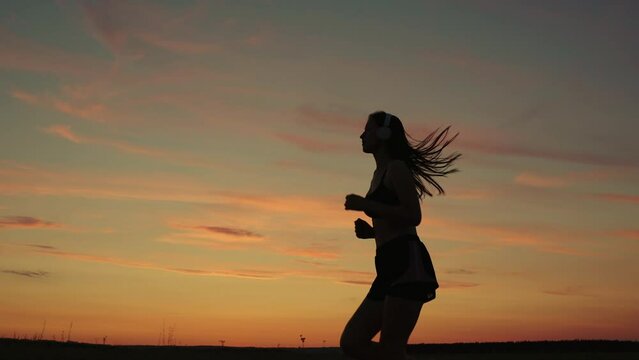Training run. Silhouette, beautiful girl in musical headphones, is engaged in fitness, runs in evening. Free young woman runs in summer in park at sunset, listens to music on headphones. Teen runner