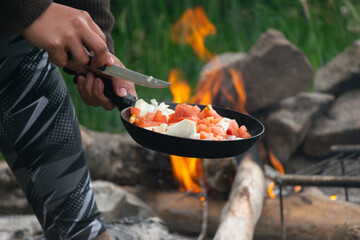 una persona cocinando verduras en una fogata con rocas . una persona acampando y cocinando en un sarten  - obrazy, fototapety, plakaty