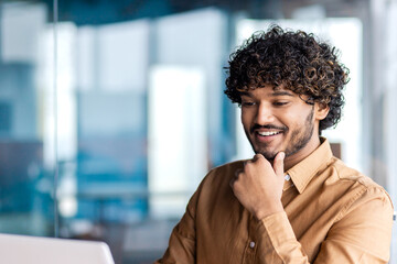 Young successful businessman close up inside office, satisfied man smiling looking at laptop...