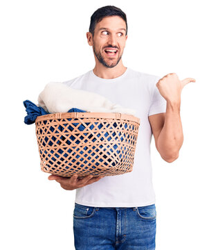 Young handsome man holding laundry basket pointing thumb up to the side smiling happy with open mouth