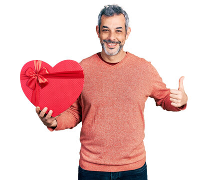 Middle age hispanic with grey hair holding valentine gift smiling happy and positive, thumb up doing excellent and approval sign