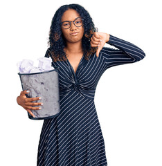 Young african american woman holding paper bin full of crumpled papers with angry face, negative sign showing dislike with thumbs down, rejection concept