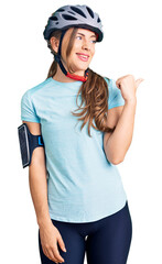 Beautiful caucasian young woman wearing bike helmet smiling with happy face looking and pointing to the side with thumb up.