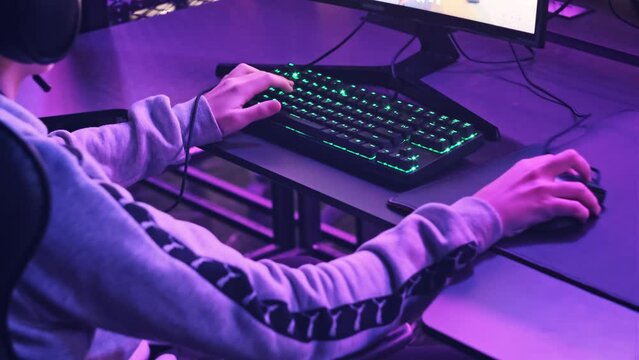 Young gamer sits in a professional gaming chair and plays video games at E-sport club. Boy in a headset at the computer club with neon lights focuses on a video game. Teenager in a gaming club, hobby