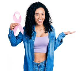 Young hispanic woman with curly hair holding pink cancer ribbon celebrating achievement with happy smile and winner expression with raised hand
