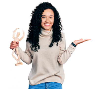 Young hispanic woman with curly hair holding small wooden manikin smiling cheerful presenting and pointing with palm of hand looking at the camera.