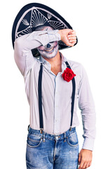 Man wearing day of the dead costume over background covering eyes with arm smiling cheerful and funny. blind concept.