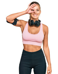 Young blonde woman wearing gym clothes and using headphones doing ok gesture with hand smiling, eye looking through fingers with happy face.