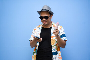 Asian tourist man wearing a beach shirt holding mobile phone with blank screen and banknotes, advertising holiday package promos isolated over light blue background. vacation concept