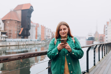 Fototapeta premium Attractive young female tourist is exploring new city. Gdansk old town and famous Zuraw crane, A happy beautiful 30s woman with smart phone is standing near Motlawa river