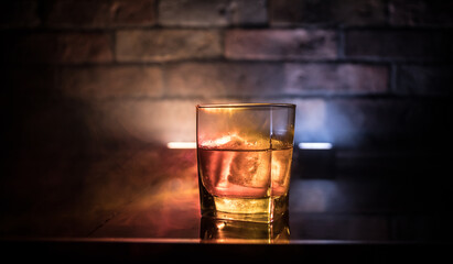 Whiskey in fire concept. Glass of whiskey and ice on wooden surface with color light and fog on...