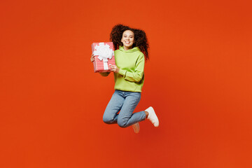 Full body young fun woman of African American ethnicity she wear green hoody casual clothes jump high hold present box with gift ribbon bow isolated on plain red orange background. Lifestyle concept.