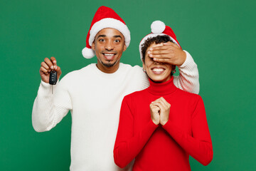 Merry young couple friends man woman wear casual clothes Santa hat posing hold car key fob keyless system cover eyes isolated on plain green background. Happy New Year 2024 Christmas holiday concept.