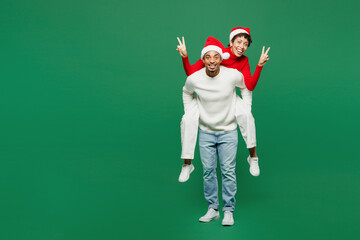 Full body merry young couple man woman wearing red casual clothes Santa hat posing give piggyback ride to joyful, sit on back isolated on plain green background Happy New Year 2024 Christmas concept