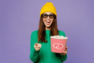 Young woman wears green sweater yellow hat casual clothes 3d glasses watch movie film hold bucket of popcorn in cinema point finger camera on you isolated on plain purple background studio portrait.