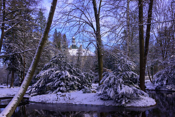 Pałac w parku zimą wśród drzew i krzewów są one pokryte warstwą śniegu, który pokrywa również ziemię. Niebo jest błękitne, lekko zachmurzone.