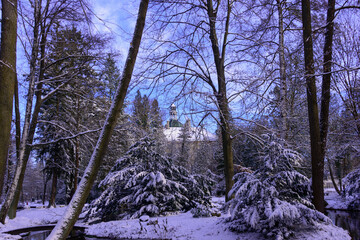 Pałac w parku zimą wśród drzew i krzewów są one pokryte warstwą śniegu, który pokrywa również ziemię. Niebo jest błękitne, lekko zachmurzone. - obrazy, fototapety, plakaty