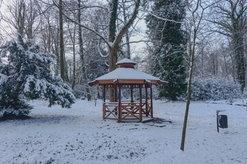 Drewniana altana w stylu japońskim w parku w Iłowej. Jest zima. Dach altany, pobliskie drzewa i ziemię pokrywa warstwa śniegu.