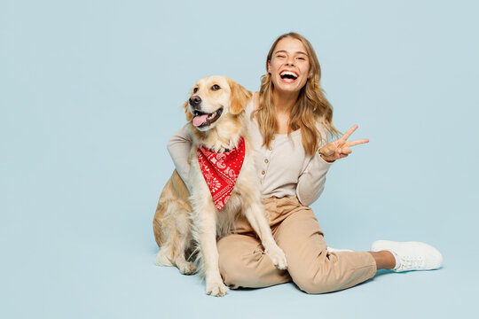 Full body young owner woman wear casual clothes sit hug cuddle embrace look at her best friend retriever dog in red bandana show v-sign isolated on plain blue background. Take care about pet concept.