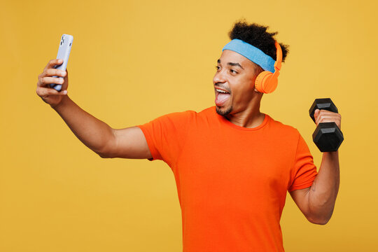 Young trainer sporty man sportsman wear orange t-shirt headphones listen music do selfie on mobile cell phone dumbbell training in in gym isolated on plain yellow background Workout sport abs concept