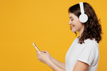 Side view young fun woman she wear white blank t-shirt casual clothes listen to music in headphones use mobile cell phone isolated on plain yellow orange background studio portrait. Lifestyle concept. - obrazy, fototapety, plakaty