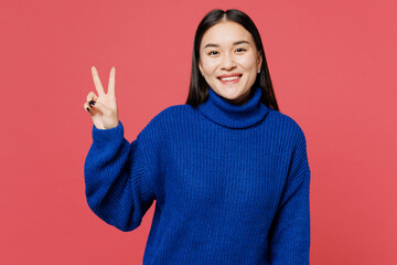 Young smiling cheerful happy woman of Asian ethnicity she wear blue sweater casual clothes showing victory sign look camera isolated on plain pastel pink background studio portrait. Lifestyle concept.