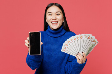 Young woman of Asian ethnicity wears blue sweater casual clothes hold in hand fan of cash money in...