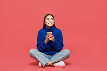 Full body young woman of Asian ethnicity she wear blue sweater casual clothes sitting hold in hand use mobile cell phone look aside isolated on plain pastel pink background studio. Lifestyle concept.