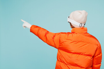 Back rear view young skier woman wearing warm padded windbreaker jacket hat ski goggles mask point finger aside travel rest spend weekend winter season in mountains isolated on plain blue background. © ViDi Studio
