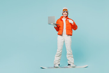 Full body young IT woman wears padded jacket hat ski goggles mask snowboarding use point on laptop...