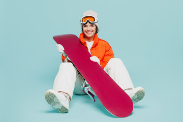 Full body young woman wears warm padded windbreaker jacket hat ski goggles mask sit with snowboard look camera travel rest spend weekend winter season in mountains isolated on plain blue background.