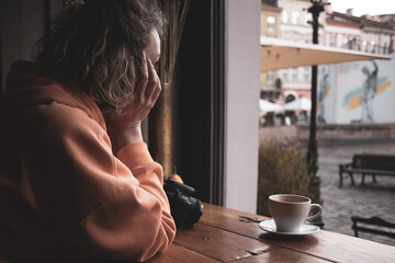 A girl in a cafe at a table. Bright casual clothes. A young woman drinks coffee. Orange sports sweater. Cup of coffee. Coffee break. Thoughtful person.