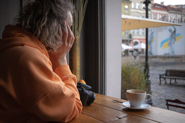 A girl in a cafe at a table. Bright casual clothes. A young woman drinks coffee. Orange sports sweater. Cup of coffee. Coffee break. Thoughtful person.