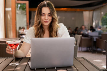 Young charming woman using laptop computer at cafe of resort hotel, drinking tropical cocktail, working typing emails browsing online