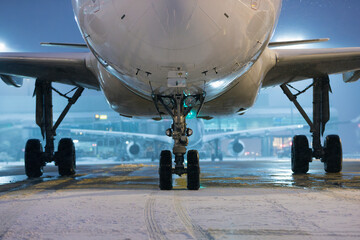 Winter frosty night at airport during snowfall. Front view of plane landing gear during taxiing to runway against airport terminal..