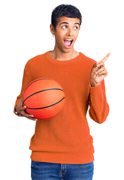 Young african amercian man holding basketball ball pointing thumb up to the side smiling happy with open mouth