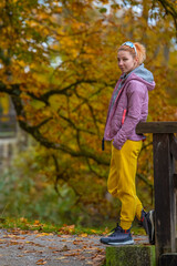 Portrait of a pretty young woman with red hair wearing a grey hoodie, pink jacket, yellow pants and hiking boots posing in a beautiful autumn park. Estonia.