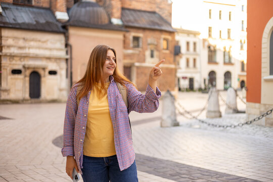Attractive young female tourist is exploring new city. Redhead girl with backpack pointing finger on city street in Krakow. Traveling in summer. St. Marys Basilica. Vacation concept