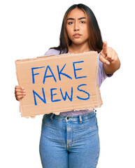 Young latin woman holding fake news banner pointing with finger to the camera and to you, confident...