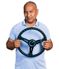 Hispanic middle age man holding steering wheel clueless and confused expression. doubt concept.