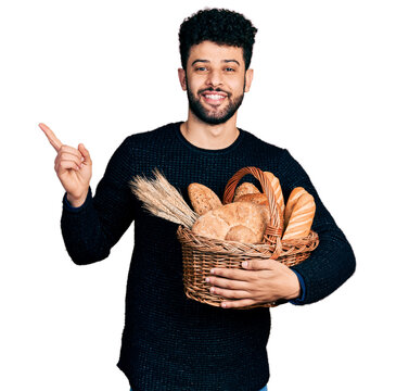 Young arab man with beard holding wicker basket with bread smiling happy pointing with hand and finger to the side