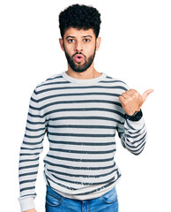 Fototapeta premium Young arab man with beard wearing casual striped sweater surprised pointing with hand finger to the side, open mouth amazed expression.