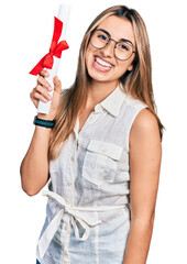 Hispanic young woman holding graduate degree diploma looking positive and happy standing and smiling with a confident smile showing teeth