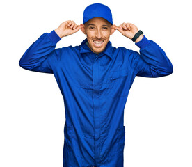 Bald man with beard wearing builder jumpsuit uniform smiling pulling ears with fingers, funny gesture. audition problem